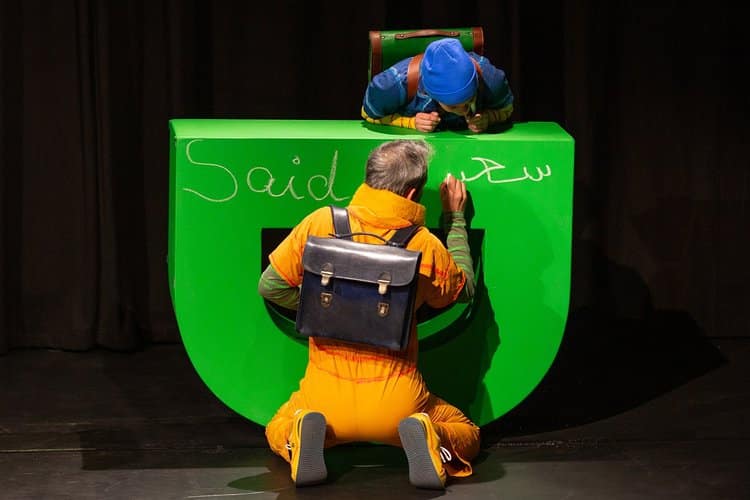 Ein Mann schreibt in arabischer Schrift auf ein grünes Bühnenteil, von oben schaut eine Frau zu