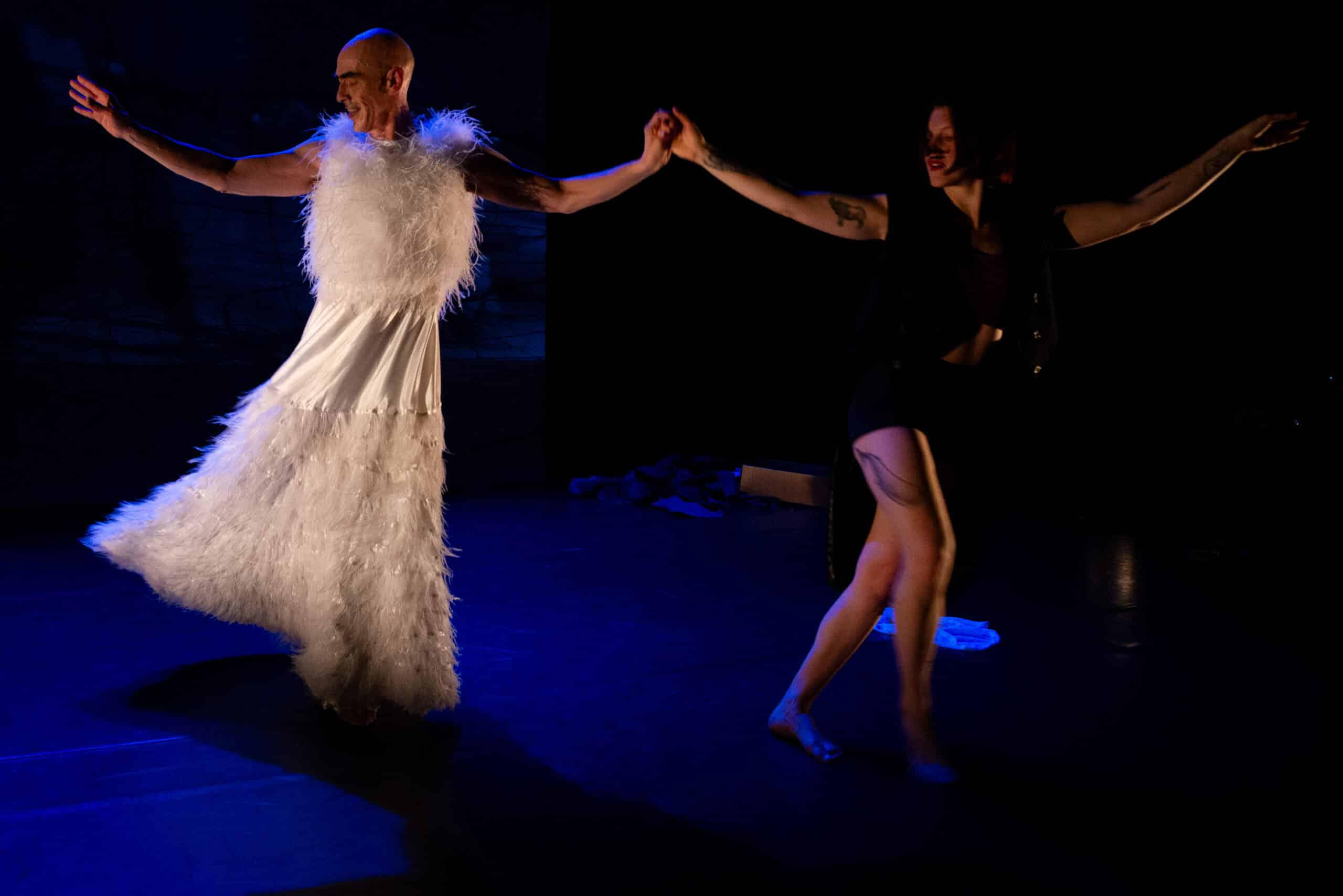 Ein Mann in weißem Kleid, eine Frau in schwarzen kurzen Hosen, sie halten sich an den Händen und tanzen