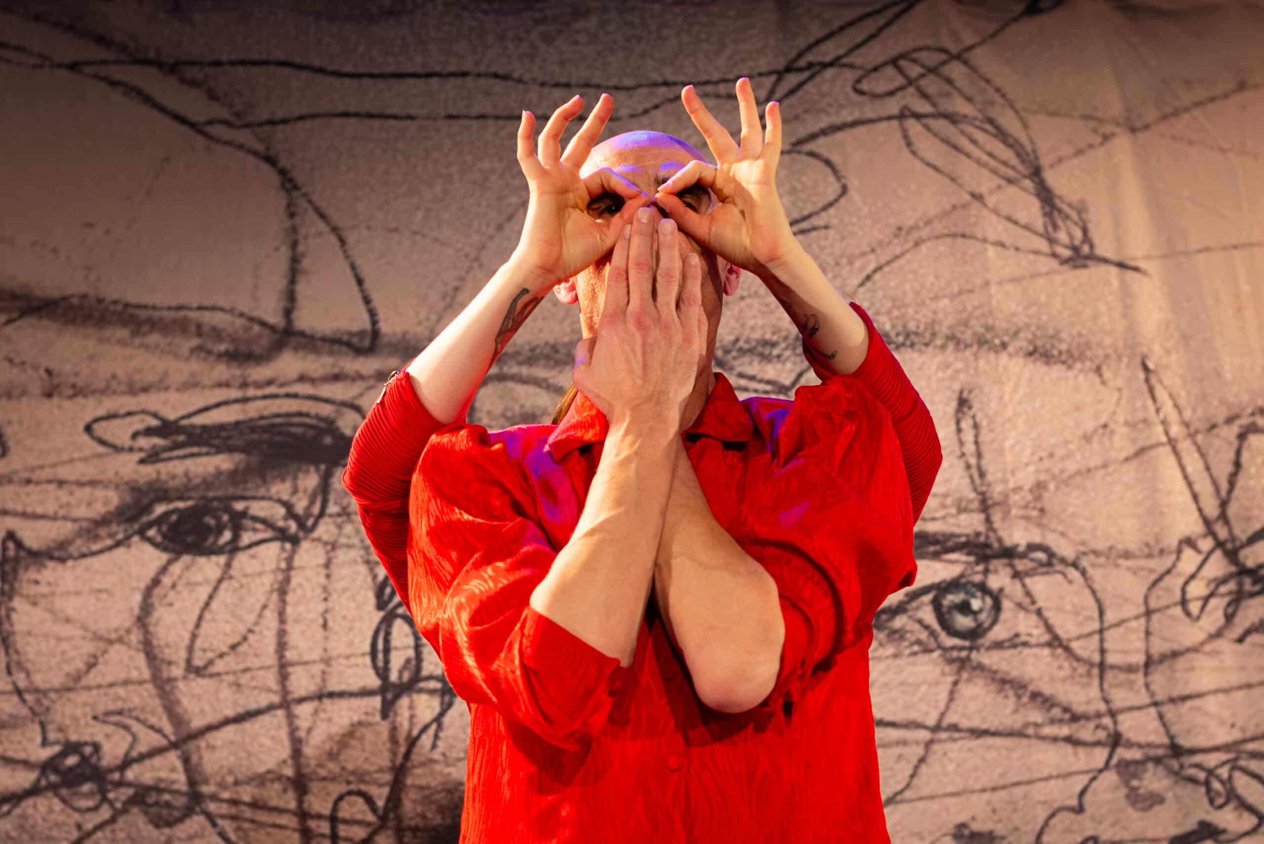 Ein Mann in rotem Hemd, er hält seine Hände vor dem Gesicht, zwei weitere Hände sind um seine Augen