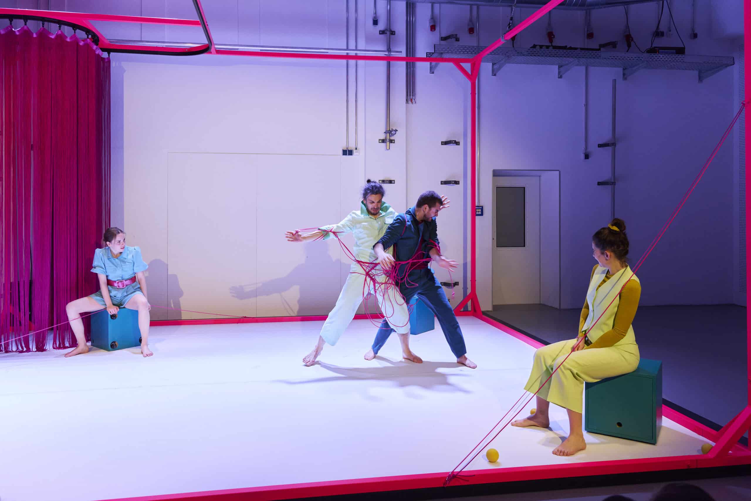 4 Menschen in einem rot umrandeten Raum, zwei sitzen abseits auf Kisten, zwei sind mit einem Faden ineinander verknotet